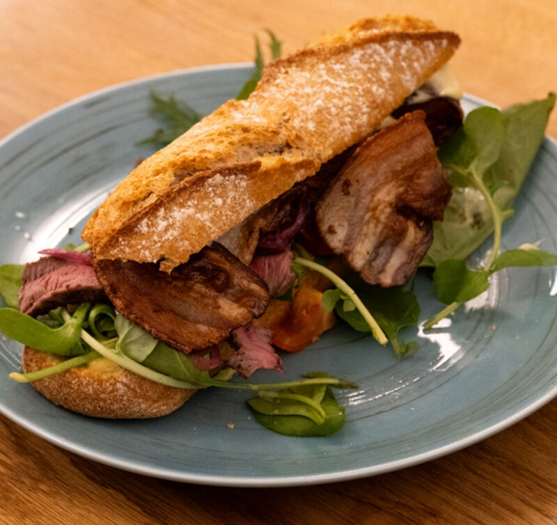 Ein aufgeschnittenes Baguettesandwich mit rosa gebratenem Fleisch und knusprigem Speck, garniert mit Rucola, auf einem blauen Teller
