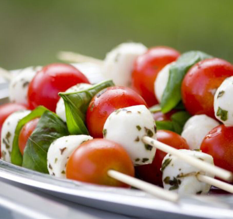 Mehrere Holzspieße mit abwechselnd aufgereihten Kirschtomaten, Mozzarellakugeln und Basilikumblättern auf einer Platte