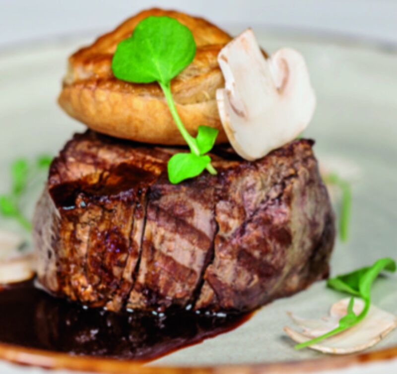 Ein angeschnittenes Stück gebratenes Rindfleisch in dunkler Sauce, obenauf ein Blätterteig-Deckel, eine Champignonscheibe und ein Kressenblatt, auf einem Teller